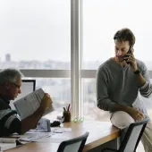 Two men in a windowed office talking; one is sitting on the desk on the phone.