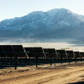 A scenic view of a solar farm with rows of solar panels in the foreground and snow-capped mountains in the background under a clear sky