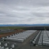 Aerial view of a large energy storage facility featuring rows of white battery storage containers and an adjacent electrical substation. The site is set in an open rural area with green fields, distant solar arrays, and a mountain range under a cloudy sky.
