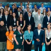 Large corporate group photo with diverse professionals wearing conference badges arranged on tiered blue seating in modern auditorium, including executive in center and attendees in business attire