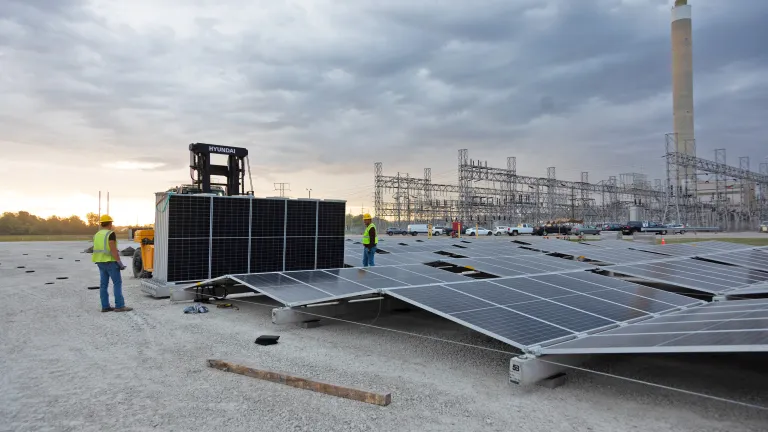 Workers installing solar panels at a power plant with a forklift and electrical infrastructure in the background under a cloudy sky.