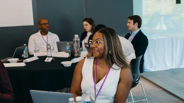 Professional woman with glasses and conference badge smiling at laptop during business meeting, with colleagues and presentation screen in background