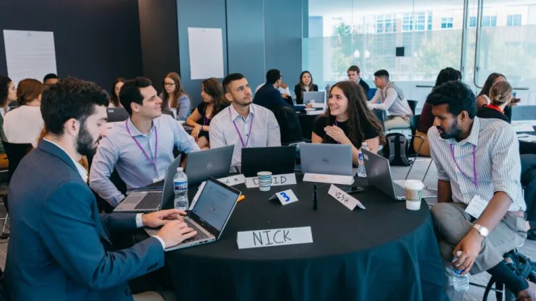 usiness workshop with professionals collaborating at tables with laptops and name tags, participants engaged in discussion in modern conference room with windows