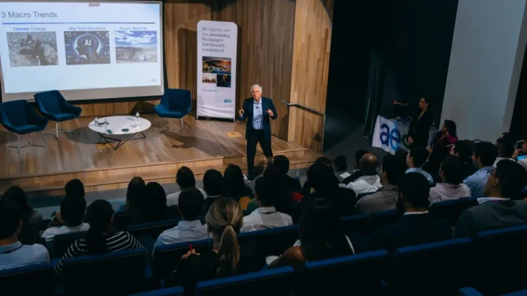 Executive presenting to large audience in auditorium with presentation screen showing macro trends including climate change and AI