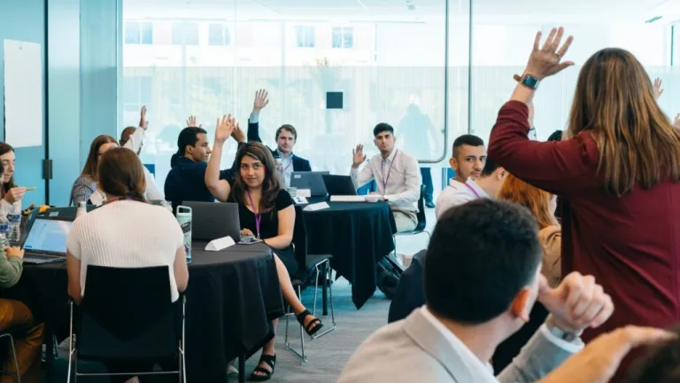 Interactive workshop session with participants raising hands while seated at tables with laptops in bright conference room
