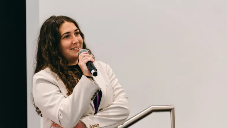 Professional woman in white blazer speaking into microphone at podium during corporate presentation or conference event
