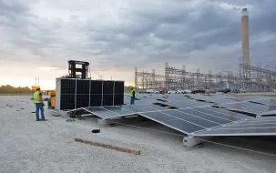 Workers installing solar panels at a power plant with a forklift and electrical infrastructure in the background under a cloudy sky.