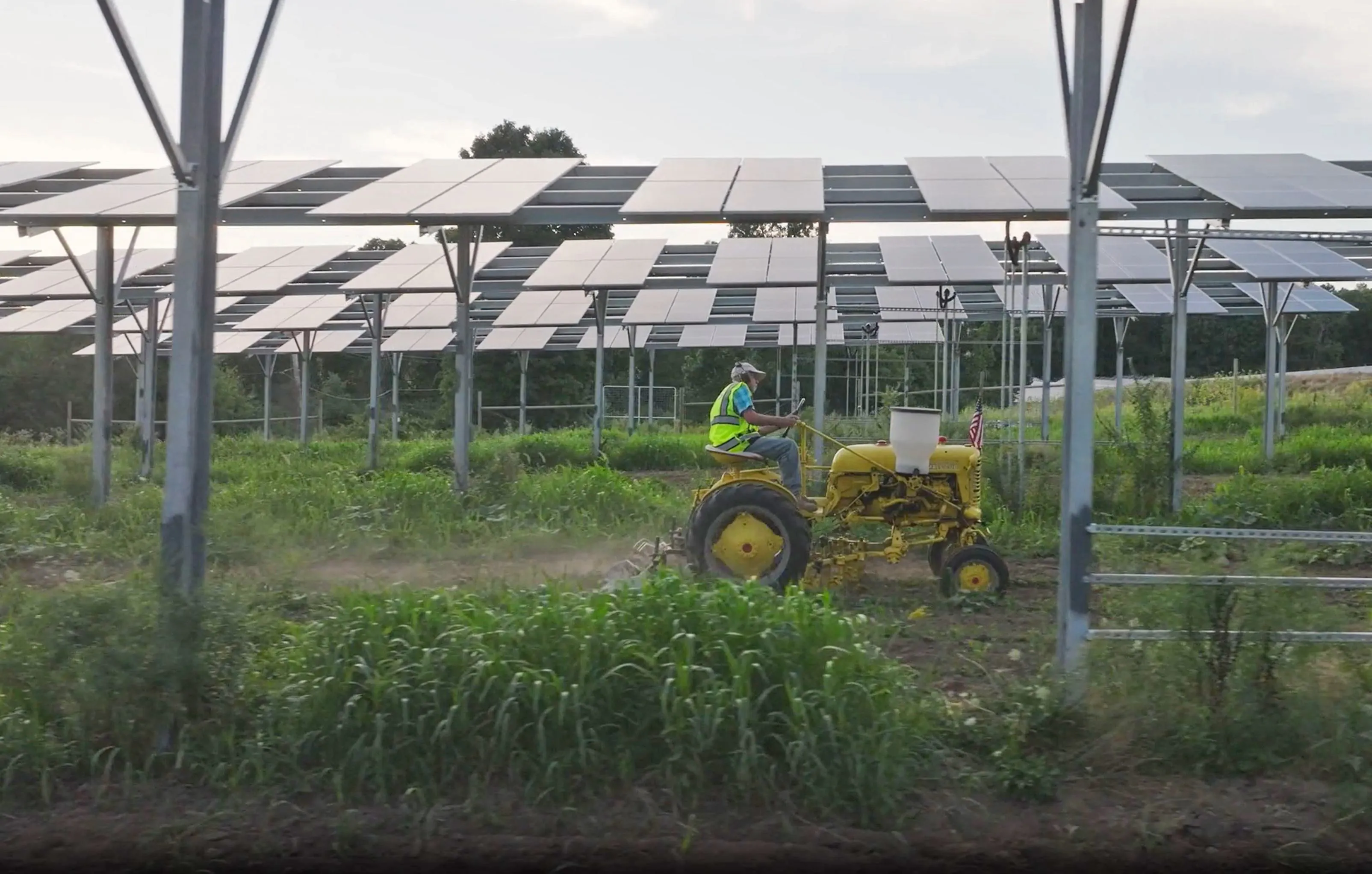 A farmer drives a yellow tractor through a green field beneath large solar panel arrays, showcasing sustainable agriculture and renewable energy integration.
