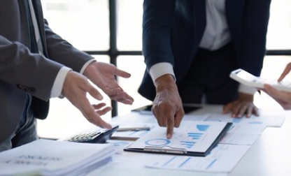 Business professionals stand around a table with documents and a tablet, discussing data shown in charts and graphs. One person points to a document with a pen, while another holds a smartphone.