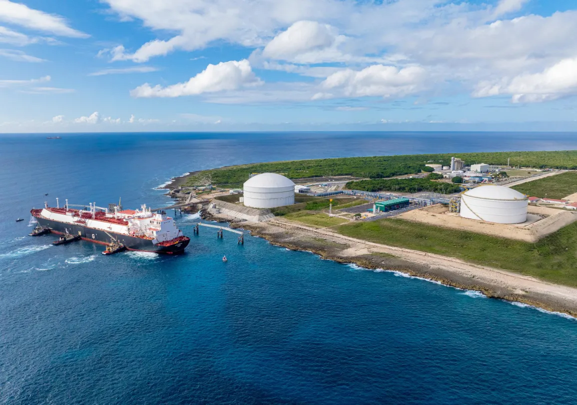 Aerial view of a coastal industrial area with two large white storage tanks near the shoreline. A large ship is docked at the pier, surrounded by clear blue water. The background shows green vegetation and a blue sky with scattered clouds.