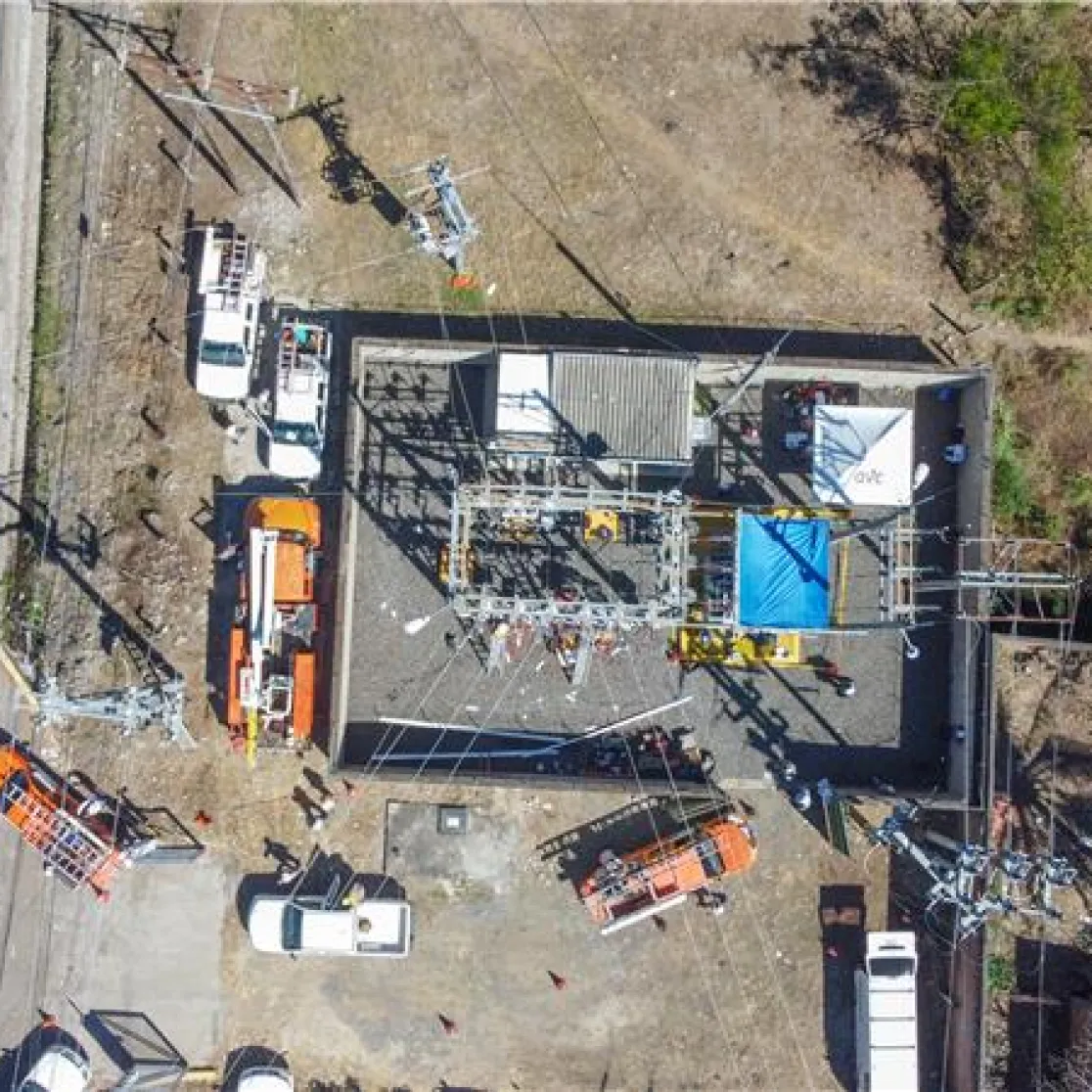 Aerial view of a power substation surrounded by several utility vehicles, including orange and white trucks, parked on a paved area. The substation is enclosed by a fence and contains electrical equipment. Nearby, there is a road on the left and grassy areas with trees on the right.