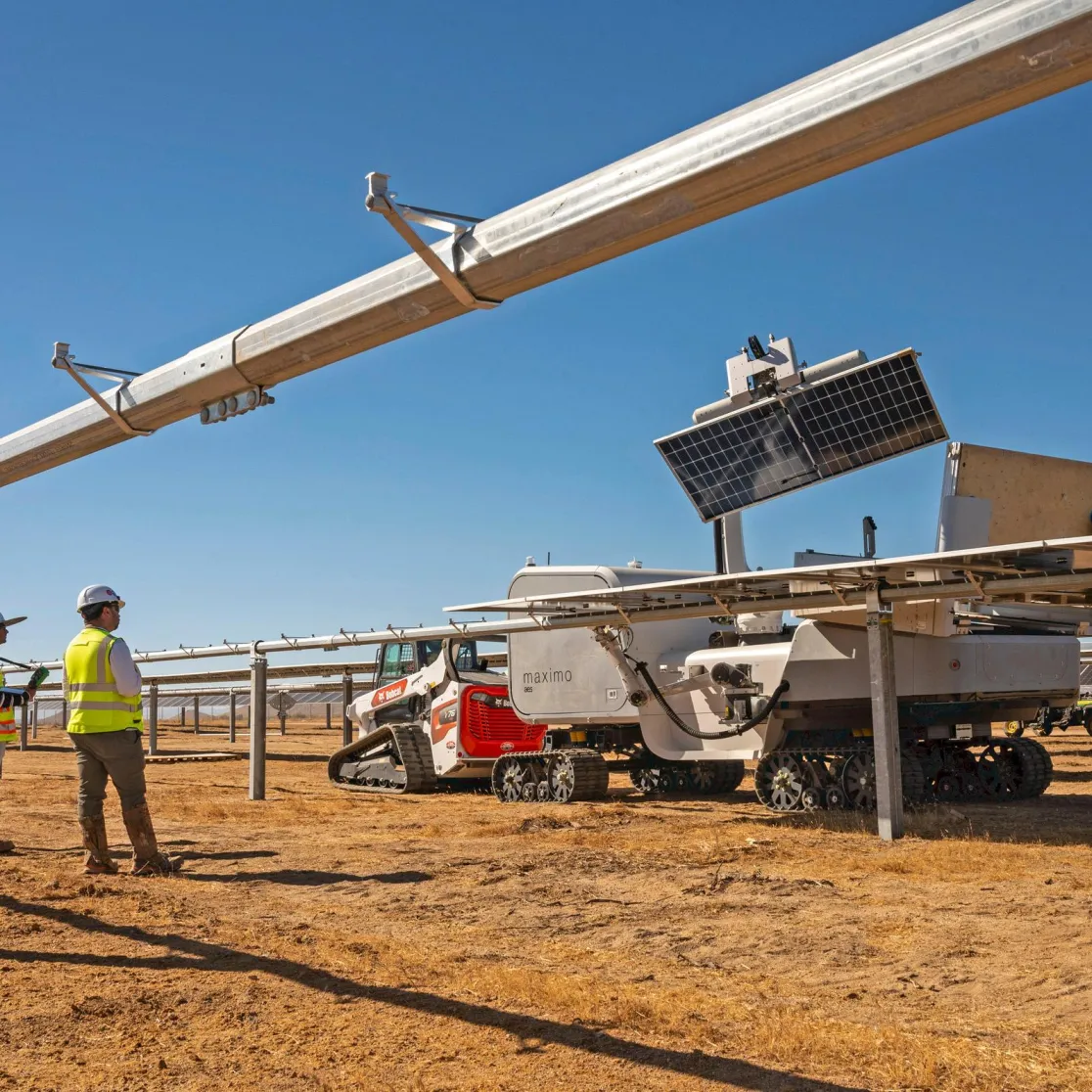 Three workers in safety gear stand beside a solar panel installation in a dry, open field. A robotic machine with tracks is positioned under the panels, likely for maintenance or cleaning. The sky is clear and blue.