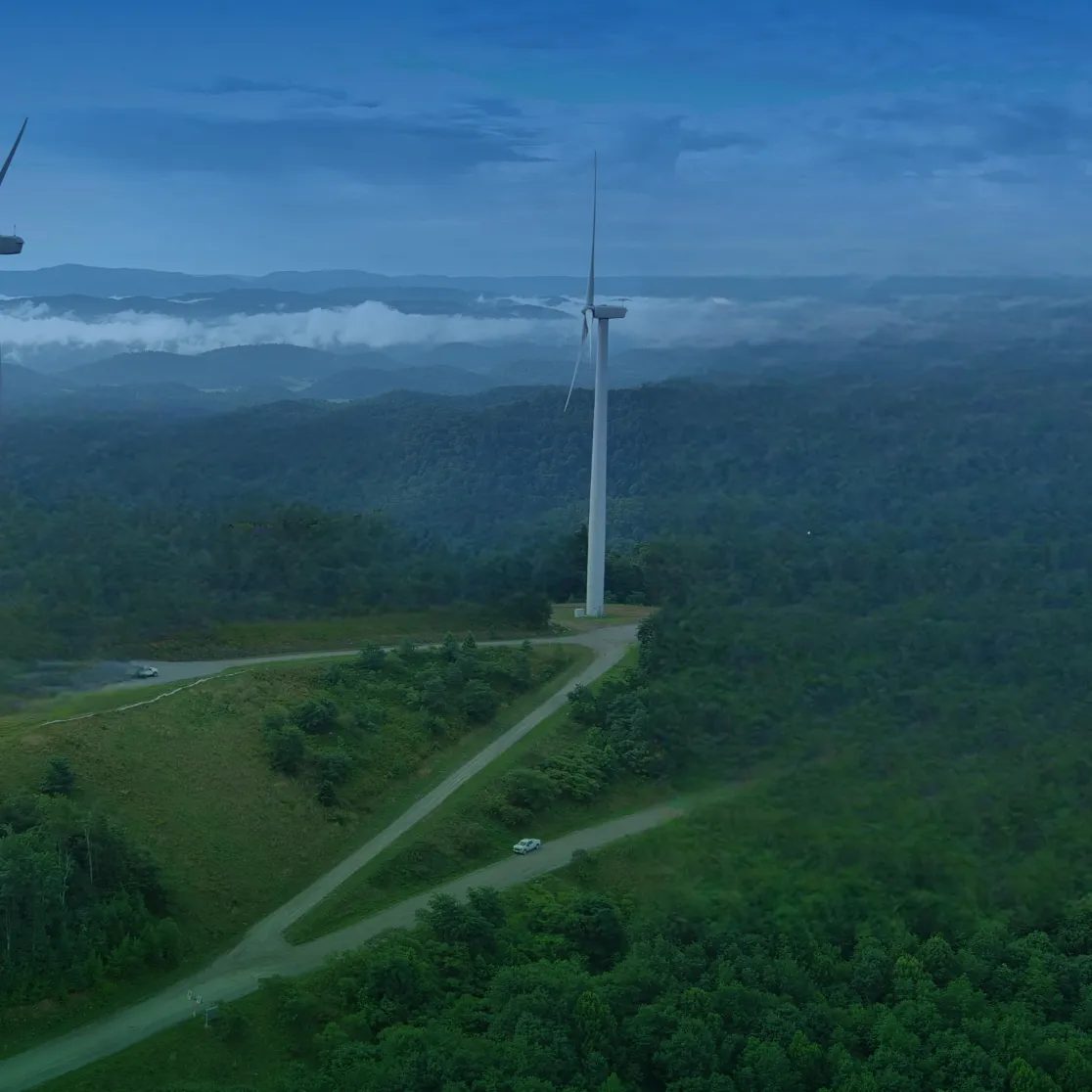 Aerial view of a lush green landscape with multiple wind turbines scattered across the hills. A winding road leads through the forested area, with a single vehicle visible on it. The sky is clear with a few clouds on the horizon.