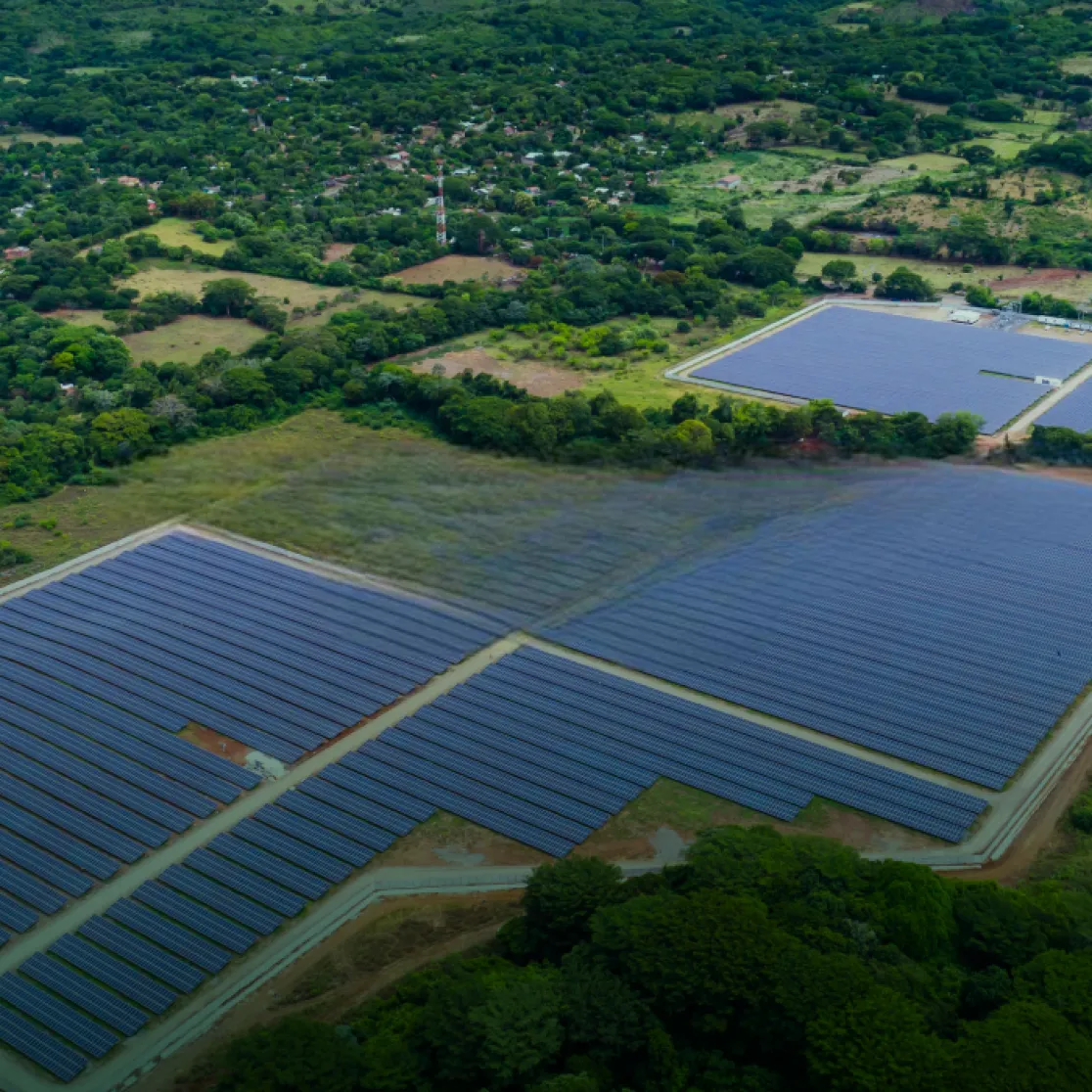 Aerial view of a large solar farm with rows of solar panels surrounded by green fields and trees. In the background, a small village is visible amidst a lush landscape.