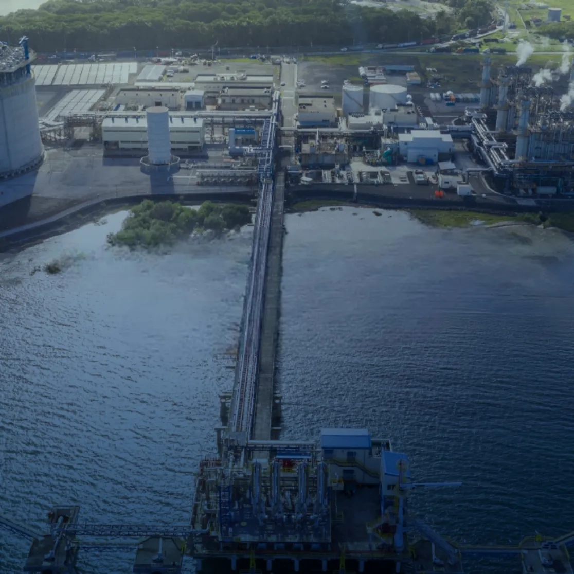 Aerial view of an industrial facility by the water. The facility includes large cylindrical tanks, various buildings, and a long pier extending into the water. Smoke is visible coming from several stacks. Greenery surrounds the area, with a river or inlet nearby.