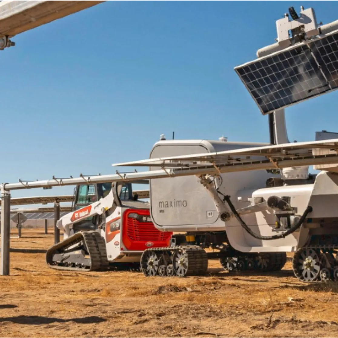 Three workers in safety gear observe a large, tracked solar panel cleaning robot labeled 'maximo' operating in a solar farm. The robot is mounted on tracks and positioned under a row of solar panels. The setting is an open, dry landscape under a clear blue sky.