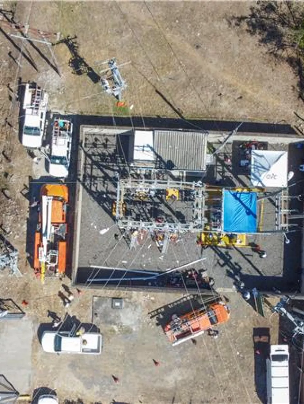Aerial view of a power substation surrounded by several utility vehicles, including orange and white trucks, parked on a paved area. The substation is enclosed by a fence and contains electrical equipment. Nearby, there is a road on the left and grassy areas with trees on the right.