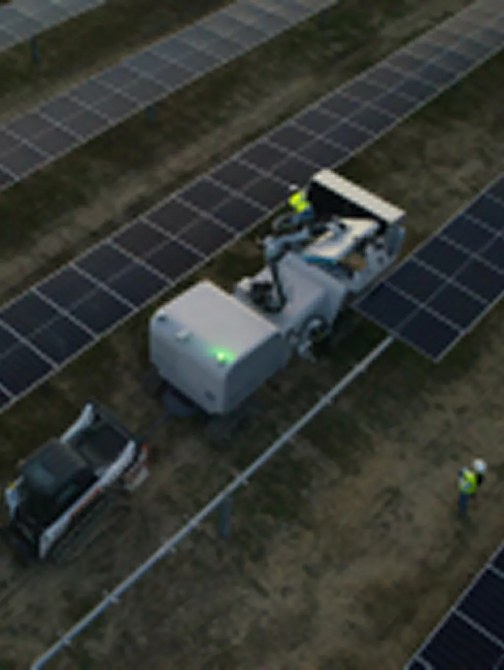 Aerial view of a solar farm with rows of solar panels. A robotic vehicle is operating between the panels, accompanied by two workers in safety vests and helmets. The scene is captured during twilight, with artificial lights visible on the vehicle.