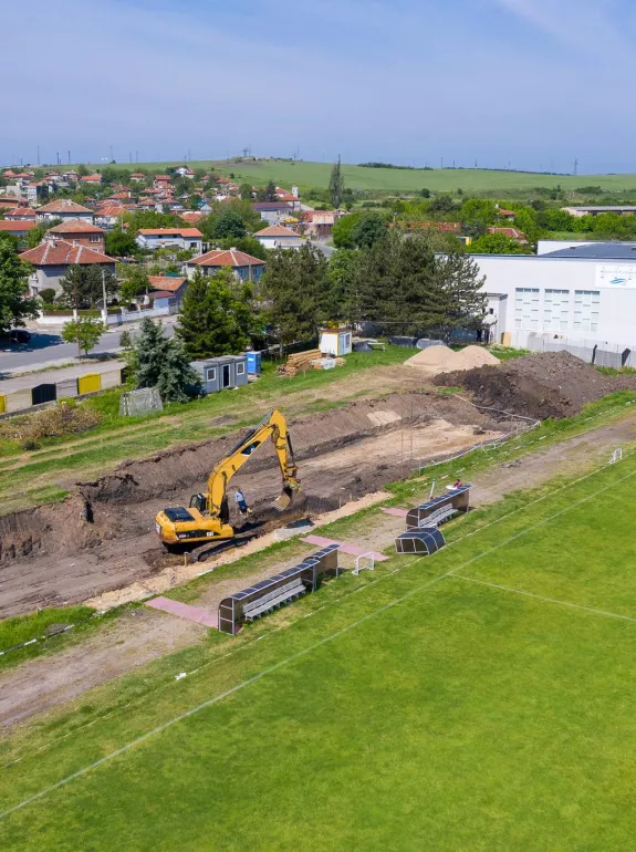 A construction site next to a sports field with a yellow excavator digging a trench. Nearby, there are benches and a small shelter. In the background, residential buildings and a large white building are visible, surrounded by trees and green hills under a blue sky.