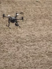 Two people in high-visibility jackets and helmets stand in a field, operating a drone. The drone is flying close to them over dry, brown grass.