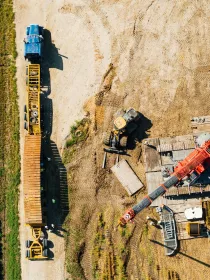 Aerial view of a construction site beside a green field. A blue truck with a long yellow trailer is parked on a dirt path. Nearby, a yellow excavator and a red crane are positioned on wooden planks. The area shows visible tire tracks and scattered construction materials.