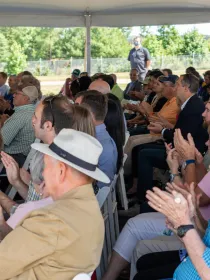 A large group of people sitting under a tent, clapping and facing forward. The audience includes men and women of various ages, some wearing hats. The setting is outdoors with trees visible in the background.