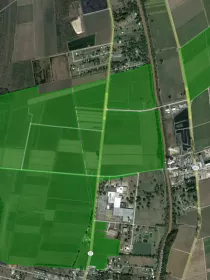 Aerial view of farmland with various green fields outlined in bright green. Roads intersect the fields, with a few buildings visible in the bottom right corner. The landscape includes patches of trees and a small body of water.