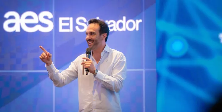 A person in a white shirt holds a microphone and gestures with their hand while speaking on stage. The background displays the text 'aes El Salvador' on a large screen.