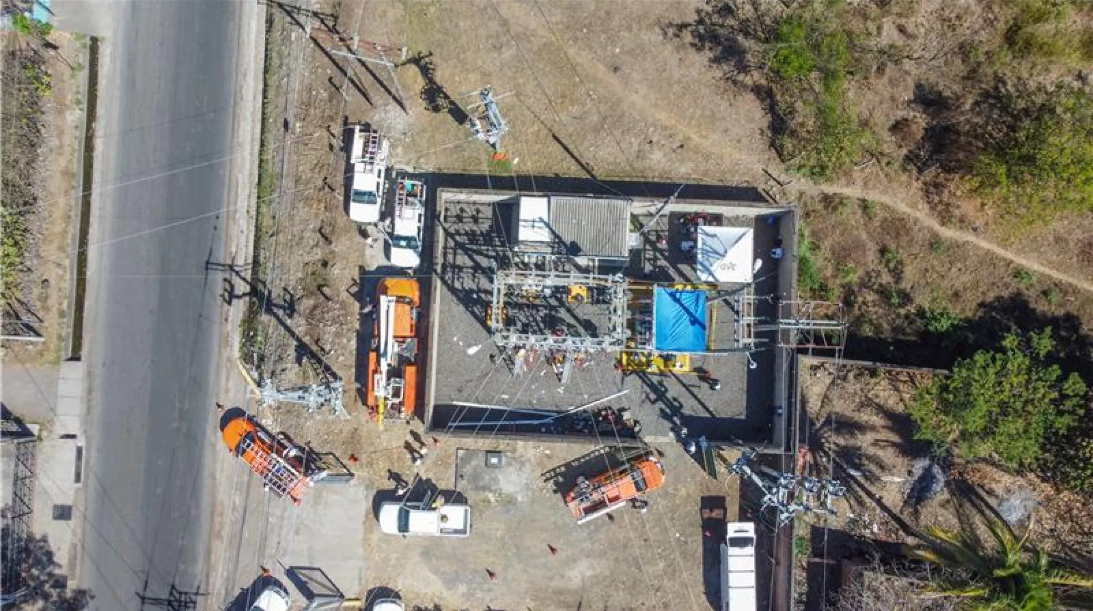 Aerial view of a power substation surrounded by several utility vehicles, including orange and white trucks, parked on a paved area. The substation is enclosed by a fence and contains electrical equipment. Nearby, there is a road on the left and grassy areas with trees on the right.