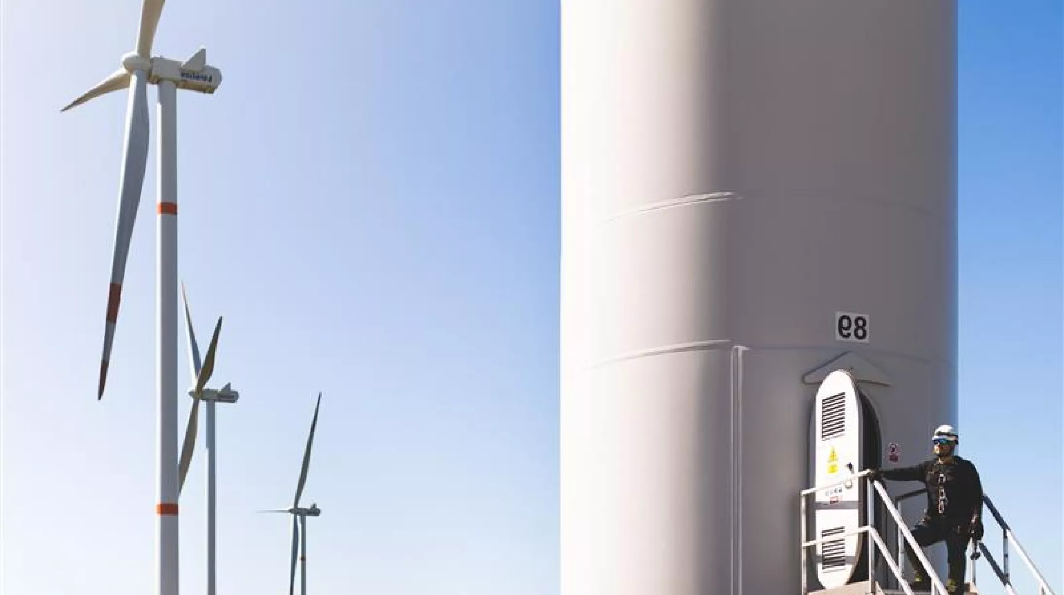 A person wearing safety gear stands on a platform at the base of a large wind turbine with the label 'e8'. Several wind turbines are visible in the background against a clear blue sky.
