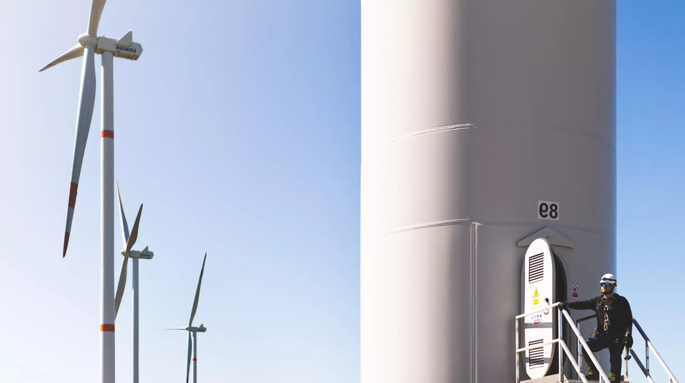 A person wearing safety gear stands on a platform attached to a large wind turbine tower labeled 'e8'. In the background, three more wind turbines are visible against a clear blue sky.