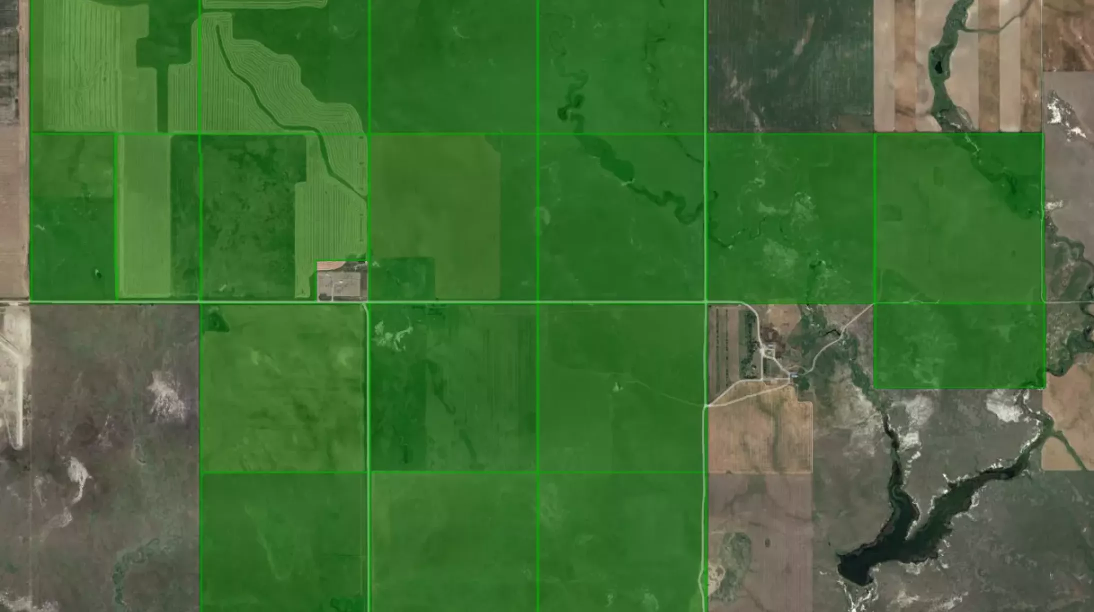 Aerial view of farmland divided into rectangular plots, highlighted in green. The plots contain various patterns of cultivation, surrounded by uncultivated land and a river running through the landscape.