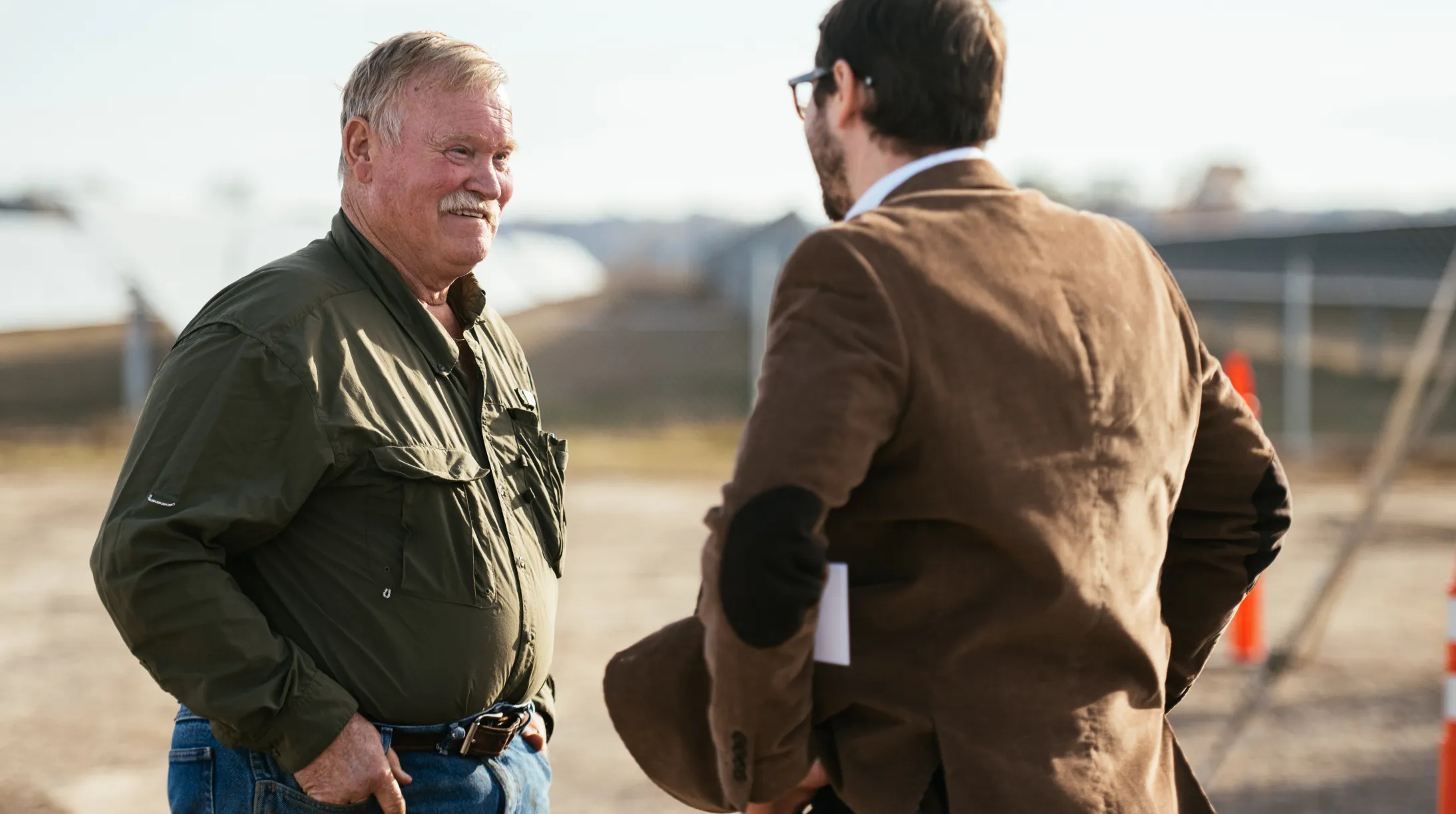 Two men are engaged in conversation outdoors. One man, wearing an olive green shirt and jeans, faces the camera. The other man, in a brown jacket with elbow patches, has his back to the camera. They are standing near a fenced area with blurred structures in the background.