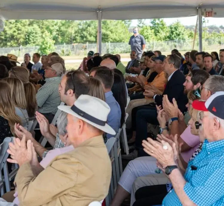 A large group of people seated under a tent, clapping and facing forward. Some individuals wear hats and sunglasses. Trees and a fence are visible in the background.