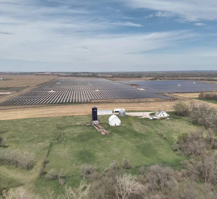 Aerial view of a large solar farm with rows of solar panels extending across a vast field. In the foreground, there is a farm with a white barn, silo, and a house surrounded by green grass and trees. The sky is partly cloudy.