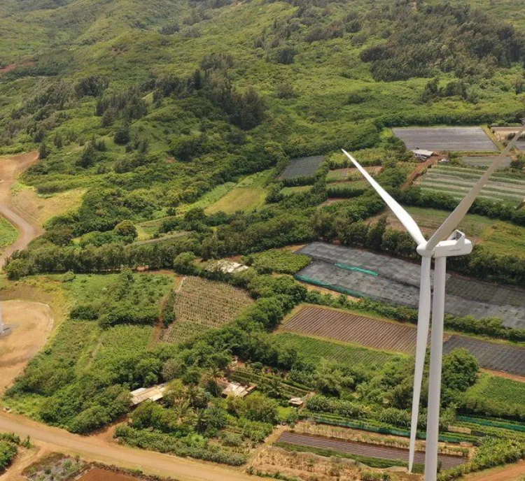 Aerial view of two large wind turbines situated among lush green farmland and hills. The landscape includes various agricultural plots with different shades of green, indicating diverse crops. Dirt roads wind through the area, and dense forested hills are visible in the background.