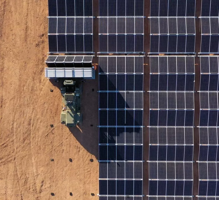 Aerial view of a large solar panel array on the right side, with a tractor and trailer carrying additional panels on the left, set on a dirt surface.