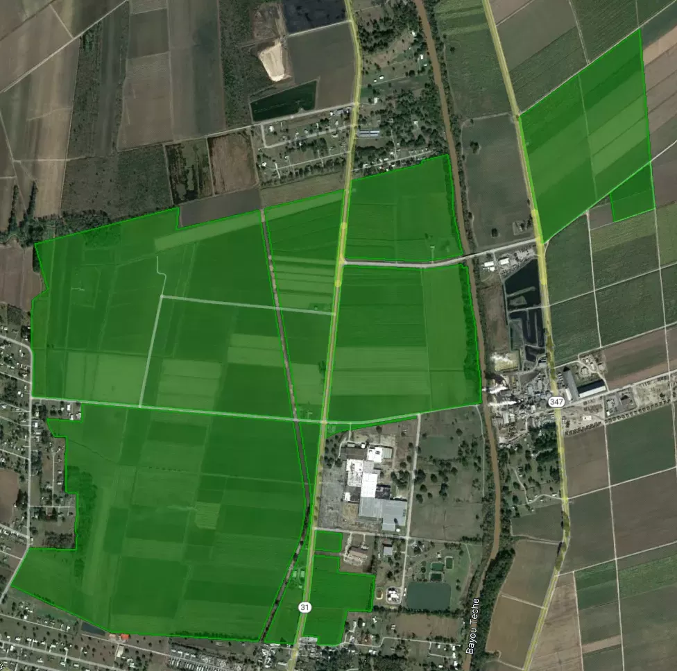 Aerial view of farmland with various green fields outlined in bright green. Roads intersect the fields, with a few buildings visible in the bottom right corner. The landscape includes patches of trees and a small body of water.