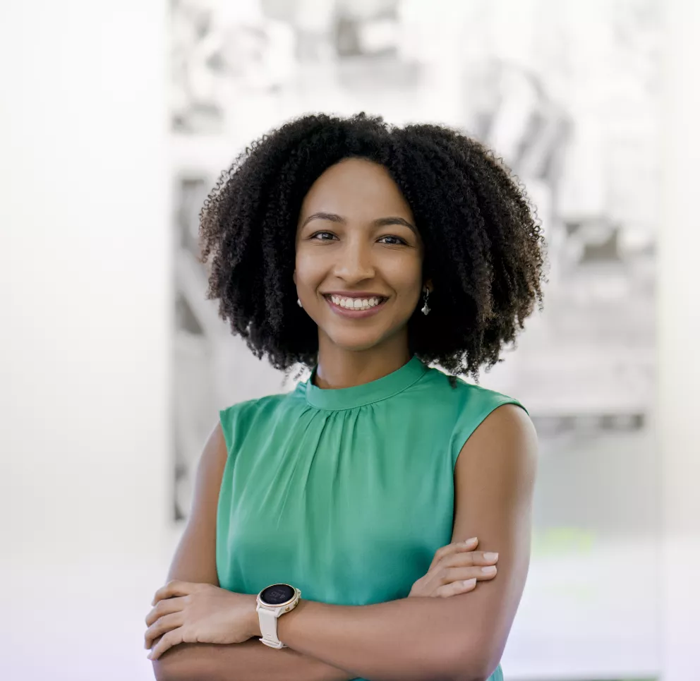 A person with curly hair smiles, standing with arms crossed wearing a green sleeveless top and a watch. The background is blurred.
