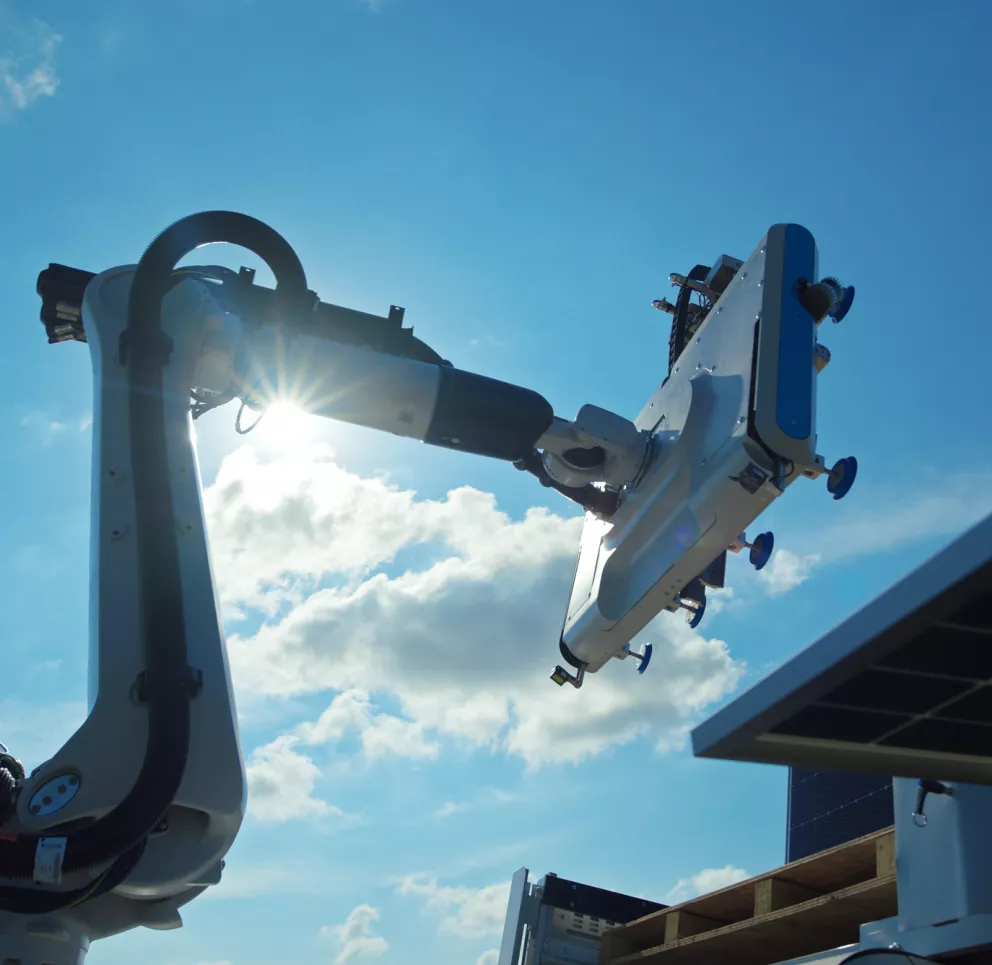 A robotic arm equipped with a large panel is positioned against a bright blue sky with scattered clouds. Sunlight shines through the arm's joints, creating a lens flare. Part of a solar panel is visible in the lower right corner.