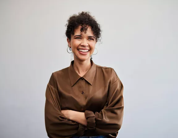 A person with curly hair smiling and crossing their arms, wearing a brown button-up shirt against a plain background.