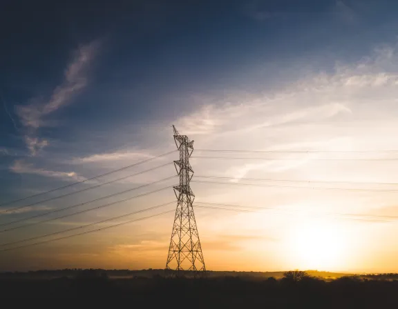 A tall electricity pylon stands against a sunset sky with scattered clouds. The sun is setting on the horizon, casting a warm golden light over the landscape. Power lines extend from the pylon across the sky.