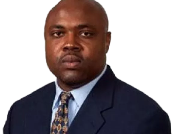 A person wearing a dark suit, light blue shirt, and patterned tie, posing against a plain background.