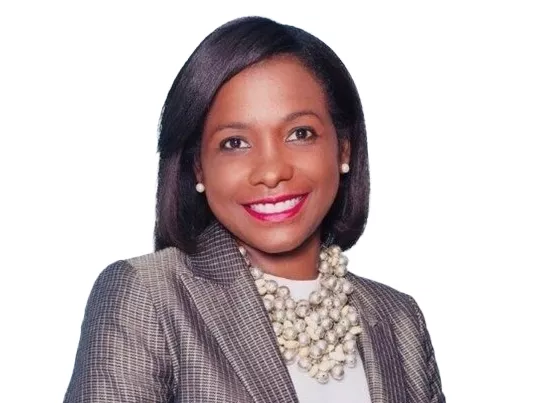 A woman with shoulder-length hair smiles, wearing a plaid blazer, white top, and a large pearl necklace. She has pearl earrings and is positioned against a plain white background.