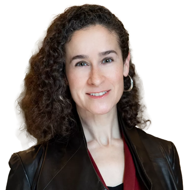 A person with curly hair, wearing a leather jacket and hoop earrings, smiles at the camera. The background is softly blurred with green foliage visible.