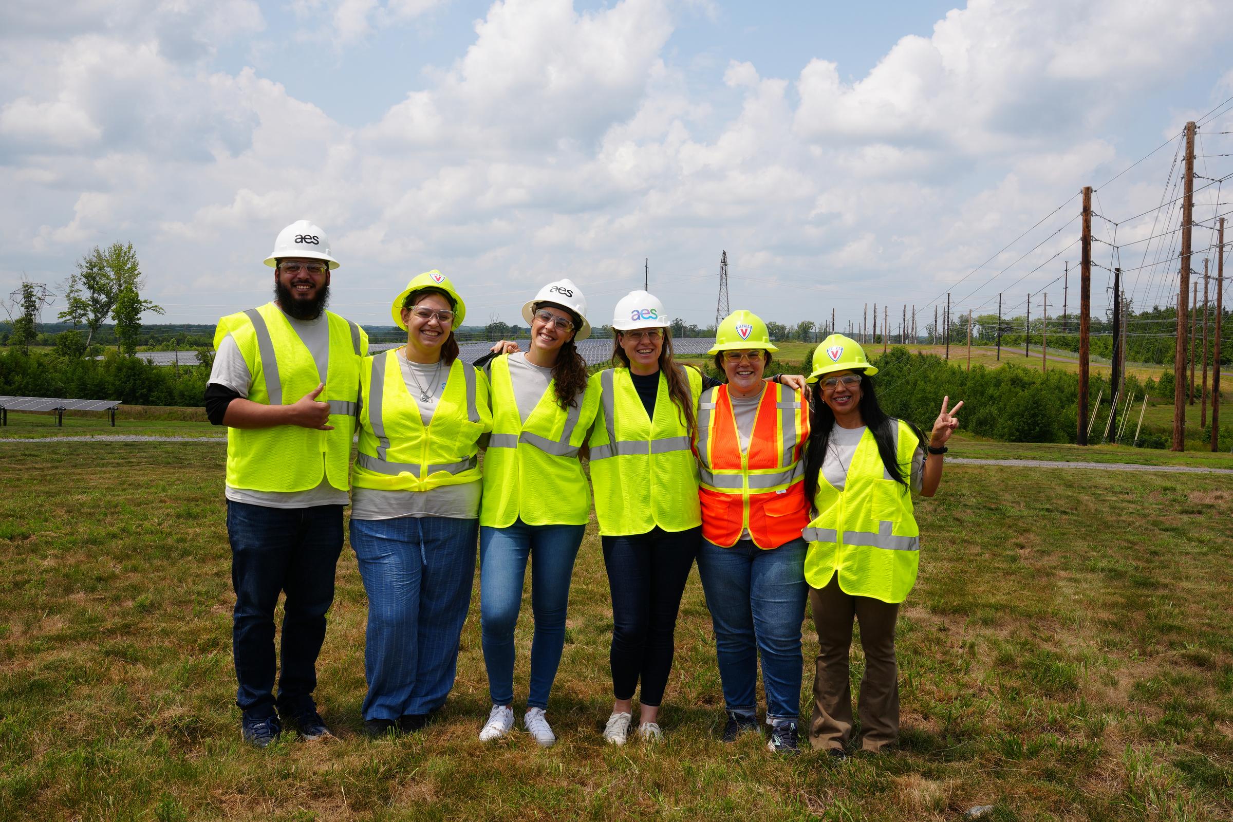 Six people wearing high-visibility vests and hard hats stand together smiling in an open grassy area with solar panels and power lines in the background. The sky is partly cloudy.