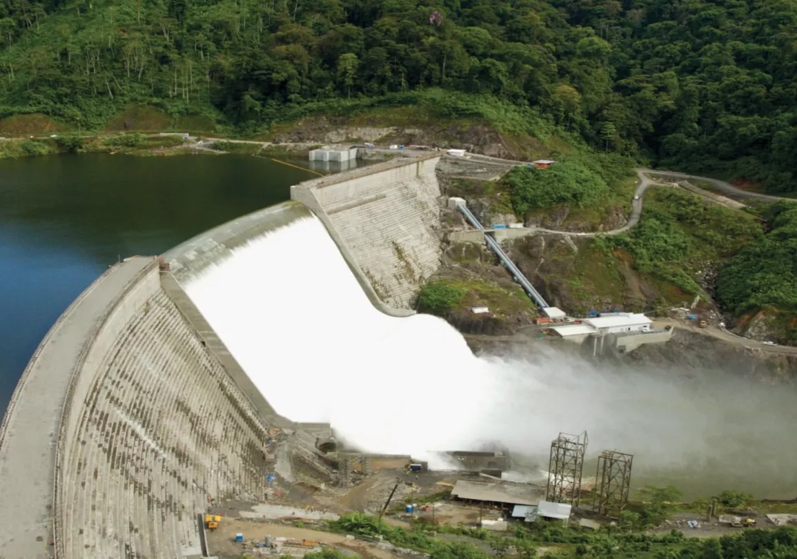 Aerial view of a large dam with water cascading over its spillway, surrounded by dense green forest. The dam structure is concrete, with water flowing into a river below. Roads and small buildings are visible near the dam, along with mist rising from the water.