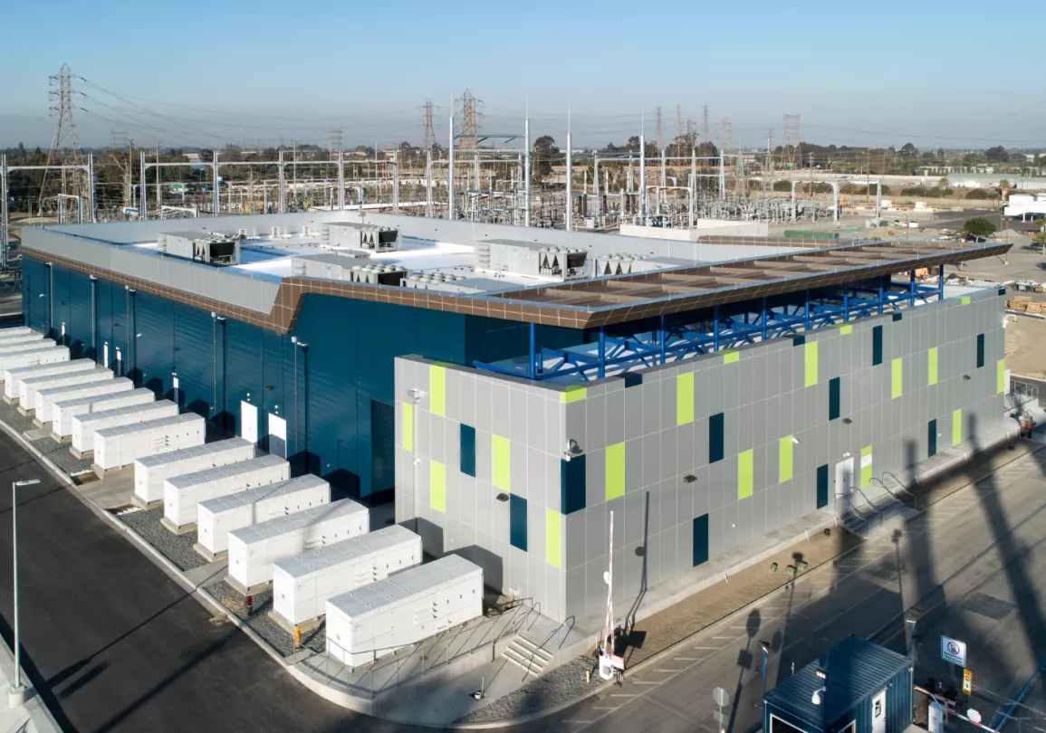 Aerial view of a large industrial facility with a modern, rectangular building featuring blue and gray panels with green accents. The building is surrounded by numerous white utility boxes and power lines. The area is paved with roads and pathways, and there are trees and other industrial structures in the background.