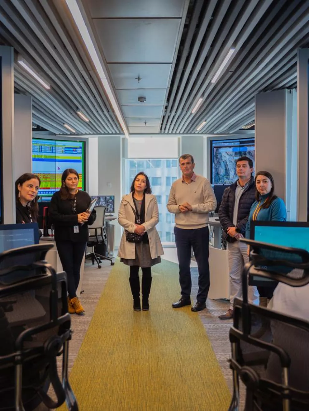 A group of people stands in a modern control room with large display screens showing graphs and data. Two individuals sit at desks with monitors in the foreground. The room has a high-tech atmosphere with multiple screens and a structured ceiling.