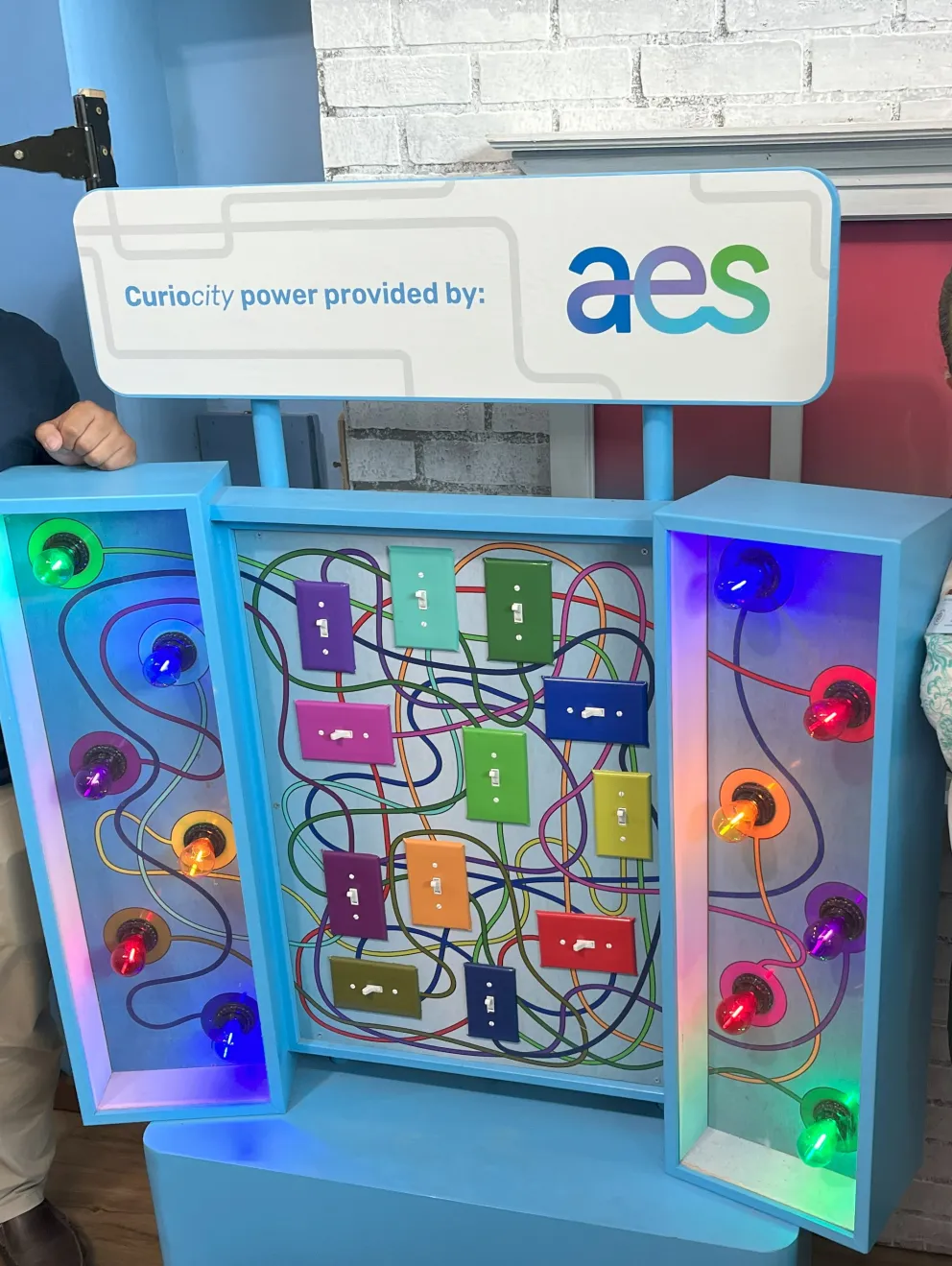 Two people stand beside a colorful interactive display with various switches and lights. The display is labeled 'Curiosity power provided by: aes'. The background features a brick wall and a wooden floor.